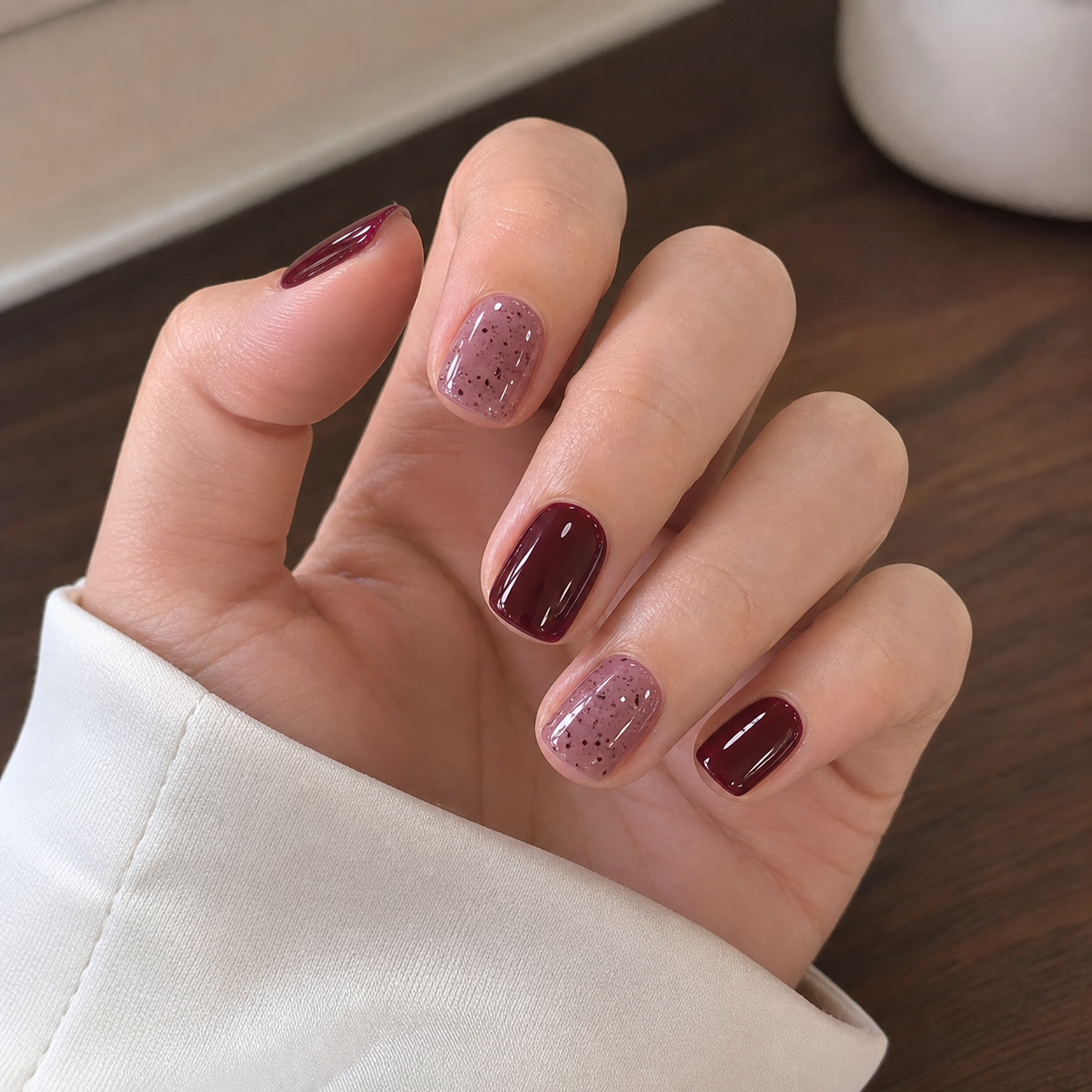 Mãos com unhas curtas e quadradas pintadas alternadamente em vermelho bordô profundo e malva translúcido com pequenos pontos escuros e acabamento brilhante.
