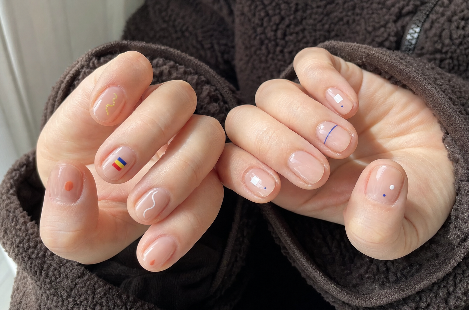 Unhas curtas e redondas com base nude translúcida e detalhes geométricos minimalistas em branco, vermelho, azul e amarelo.
