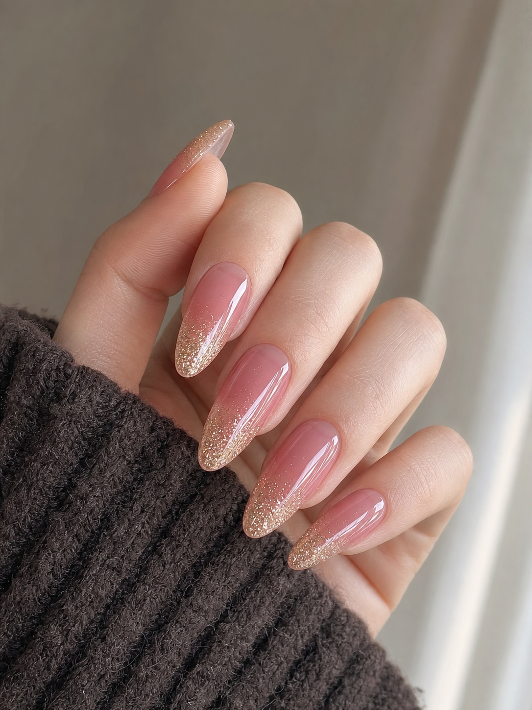 Manucure sur ongles longs en forme d'amande avec base rose jelly translucide, dégradé de paillettes dorées sur les pointes et finition top coat ultra-brillante